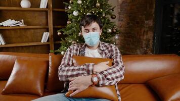 Young upset with bad mood man in protective mask is suffering from loneliness at home while sitting on couch near Christmas tree during New Year holidays. New norm, lockdown and quarantine coronavirus video