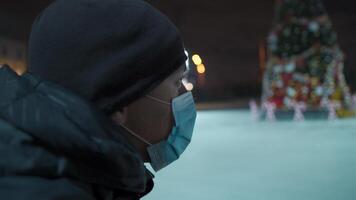 Masked male stands near skating rink arena with decorated Christmas tree empty during quarantine, coronavirus and covid pandemic in evening. Skating rink at night. An empty public ice skating area. video