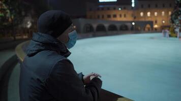 A sad man in a mask looks at an indoor ice arena with a Christmas tree in the center of the rink in the evening during lockdown, coronavirus pandemic. Empty public place for skating. video