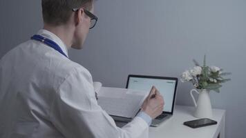 Young male physician in glasses and white lab coat working with paperwork sitting at desk operating laptop in a doctors office. Doctor reviews patients labs and input data into computer. video