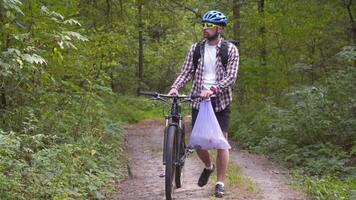 Male cyclist walking through forest with bicycle and a garbage bag collecting trash and plastic while doing sports. Bicyclist picked up trash in trash pack and walks through woods. Consciousness. video