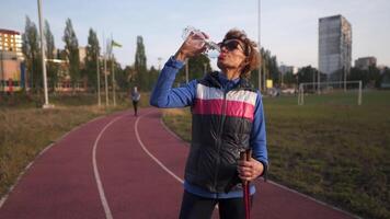 Happy senior woman drinks water from plastic bottle while training Nordic walking in city stadium. Tired elderly female refreshing herself after physical activity outdoors with Nordic walking sticks. video