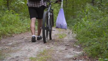 Male cyclist walking through forest with bicycle and a garbage bag collecting trash and plastic while doing sports. Bicyclist picked up trash in trash pack and walks through woods. Consciousness. video