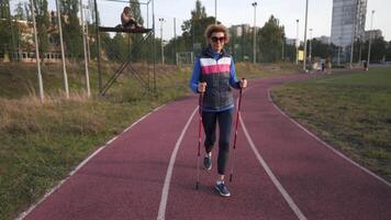 Senior woman training with walking poles at stadium. Mature female do Nordic walking exercises at running track. Healthy lifestyle concept. Active feminine exercising with trekking sticks in town. video