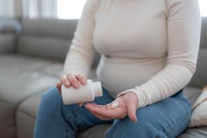 Unrecognizable pregnant woman holding jar with pills taking vitamins treating sickness during pregnancy sitting on sofa. Close up photo