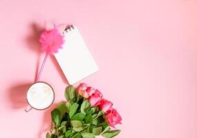 The cup of hot coffee, empty notepad with funny pen and bouquet of fresh roses on soft pink background. photo