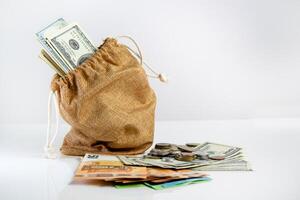 A brown burlap sack filled with US dollar bills sits on a white table next to scattered coins and bills photo