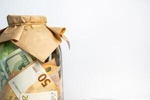 A glass jar filled with dollar and euro bills is covered with brown paper and a rubber band. The jar is on a white background photo