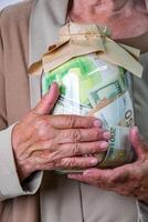 An elderly persons hands holding a glass jar filled with various bills, wrapped in a paper bag photo