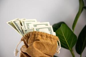 A burlap bag with a drawstring sits on a white surface, filled with US dollar bills that peek out of the top photo