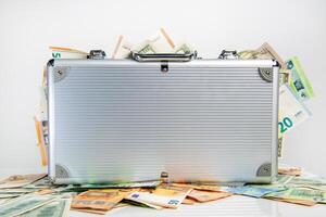 A silver suitcase is sitting on a white surface surrounded by euro and US dollar bills photo