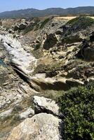 This scenic coastline features rocky cliffs, smooth erosion patterns, and patches of greenery. The area basks under bright sunlight against a backdrop of distant hills photo