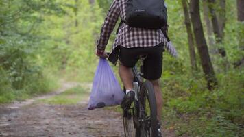 Athlete cycling riding through forest with garbage bag on the handlebars, cleaning up garbage and plastic during a bicycle trip in the forest. Ecology theme, pollution, rubbish and plastic discarded. video