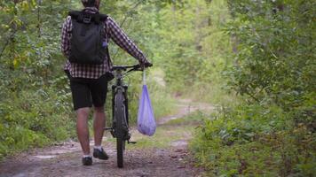 Male cyclist walking through forest with bicycle and a garbage bag collecting trash and plastic while doing sports. Bicyclist picked up trash in trash pack and walks through woods. Consciousness. video