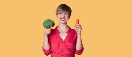 A studio shot of a smiling young woman with fresh broccoli and carrots in her hands on a yellow background. The concept of healthy eating. A place for your text. The panorama. photo