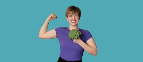 Smiling Caucasian girl with fresh broccoli in one hand and showing biceps with the other. Panoramic banner, free space. photo