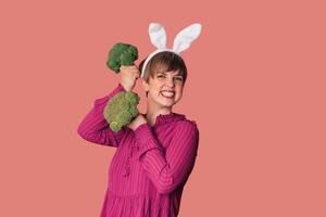 Studio shot of a young Caucasian woman in bunny ears holding fresh broccoli in her hands. healthy eating. photo