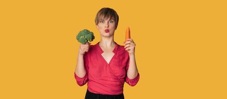un estudio Disparo de un sonriente joven mujer con Fresco brócoli y zanahorias en su manos en un amarillo antecedentes. el concepto de sano comiendo. un sitio para tu texto. el panorama. foto