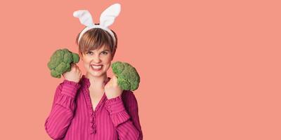 Smiling slender Caucasian girl in bunny ears with green broccoli in her hands, free space. Organic vegetables photo