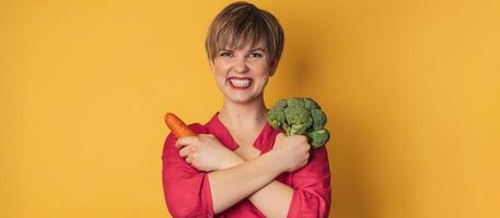 A studio shot of a smiling young woman with fresh broccoli and carrots in her hands on a yellow background. close-up.. The concept of healthy eating. A place for your text. The panorama. photo