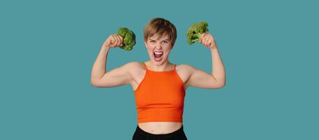 An expressive portrait of a young woman with broccoli as dumbbells on a blue background. Healthy diet, healthy lifestyle. A panoramic banner with a copy of the space. photo