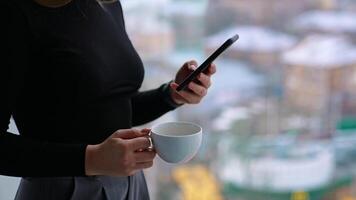Beautiful female with cup of coffee thoughtfully browsing smartphone while standing by the window video
