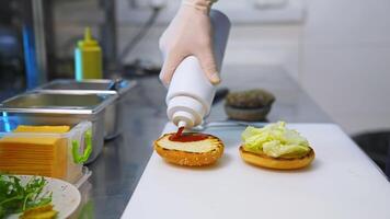 Cook adding ketchup on burger. Preparing burger on kitchen. Cook hands in white gloves preparing and making hamburger. Selective focus. video