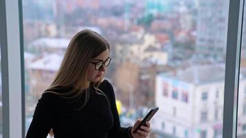 Beautiful female with cup of coffee thoughtfully browsing smartphone while standing by the window video