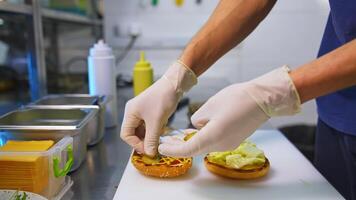 Hands in latex gloves put pickles on top of fried bun. Making burger on white table. Fast food concept . video
