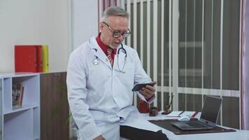 Happy satisfied mature doctor is working with patient via phone. Looking at phone in modern clinic office. Closeup. video
