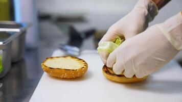 Hands in latex gloves put lettuce on top of fried bun. Making burger on white table. Fast food concept. video