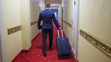 A man with a suitcase is walking down a hotel hallway. Dark red carpet in a hotel hall. View from the back on a businessman in a trip. video