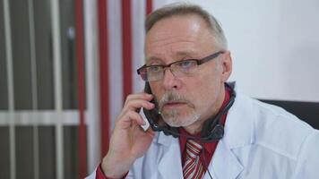 doctor sitting at desk behind his laptop and talking on mobile phone with his patient video