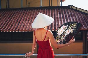 A tourist in a red dress and a traditional Vietnamese hat with a fan in her hands against the background of a tiled roof in Asia photo