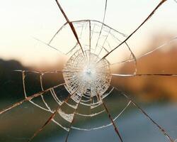 Close up of a shattered window pane with a radial crack pattern against a blurry sunset background photo