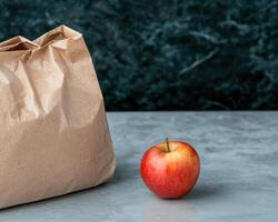 A red apple sits beside a brown paper bag on a gray surface photo