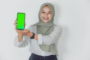 A smiling asian woman in a green hijab holds up a smartphone with a bright green screen. photo
