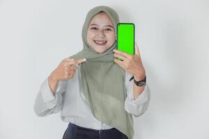 An asian woman in a green hijab smiles while showing her smartphone with a green screen. photo