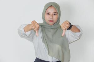 A young asian woman in a hijab expressing disapproval with thumbs down against a white background. photo