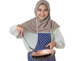 A woman in a hijab and a polka dot apron joyfully cooks with a frying pan, embodying the delight and warmth of home-cooked meals. photo