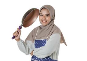 A cheerful woman in a hijab holding a frying pan and wearing a polka dot apron, ready to cook with a smile, isolated on a white background. photo