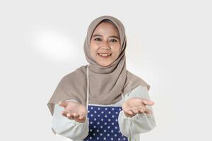 A woman wearing a gray hijab and a polka dot apron smiles and gestures with open hands, suggesting a welcoming or friendly demeanor, ready to help. photo