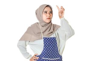 A woman wearing a hijab and a polka dot apron looks focused and contemplative, gesturing with her hand as if reaching or pointing towards something. photo