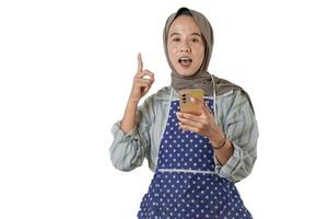 A woman wearing a hijab and apron holds a smartphone, raising her finger as if explaining or demonstrating something, with a cheerful expression. photo