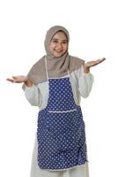 A woman wearing a gray hijab and a polka dot apron smiles and gestures with open hands, suggesting a welcoming or friendly demeanor, ready to help. photo