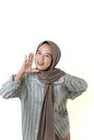 A woman wearing a hijab and striped shirt smiles while making a cheerful gesture, appearing to be calling out or speaking enthusiastically. photo