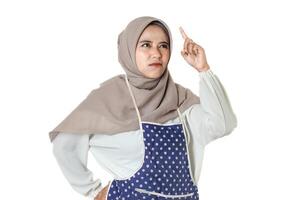A woman wearing a hijab and a polka dot apron looks focused and contemplative, gesturing with her hand as if reaching or pointing towards something. photo
