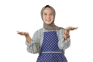 A woman wearing a gray hijab and a polka dot apron smiles and gestures with open hands, suggesting a welcoming or friendly demeanor, ready to help. photo