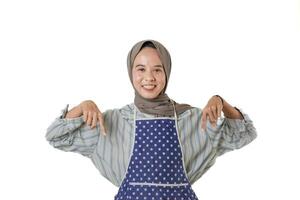 A smiling woman wearing a polka dot apron and a hijab points downward, suggesting a promotion or message below. She stands confidently against a plain background. photo