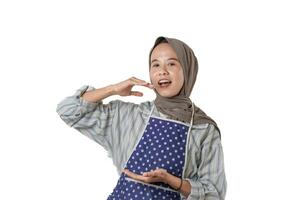 A woman wearing a hijab and an apron smiles and makes a hand gesture, possibly demonstrating something related to cooking or food presentation. photo